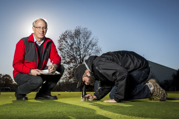 Bruce Clarke standing on annual bluegrass putting green with his former student kneeling to examine turf disease.