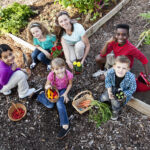 Woman (30s) with children (5 to 11 years) with flowers and vegetables in community garden.