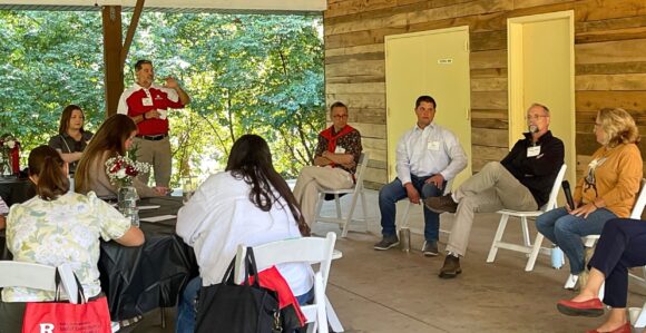 Four people sitting in a row on the right, facing several people seated at roundtables on the left. A person standing in the background is mid-speak.