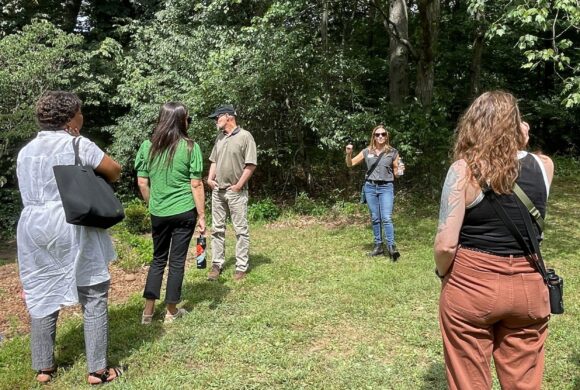 Four people standing around a woman gesturing as she speaks. They are in a grassy field. Tall, densely packed trees are in the background.