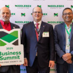 Three men are smiling in front of a Middlesex County backdrop. One holds a cardboard logo that says 2025 Business Summit.