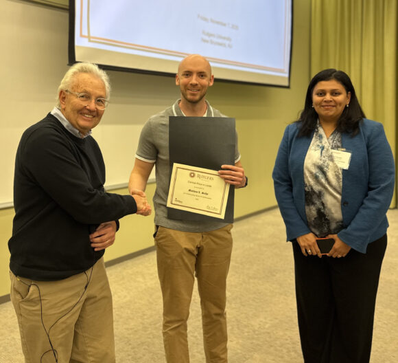This image shows a man holding an award he won. A woman stands next to him and another man shakes the award winner's hand.