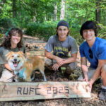 Three people and a dog pose with a plank that says RUFC 2025 in front of a boardwalk in a forest.