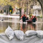 This is a very flooded street with sandbags. There is a group of people trying to help a man in an inflatable boat in the background.