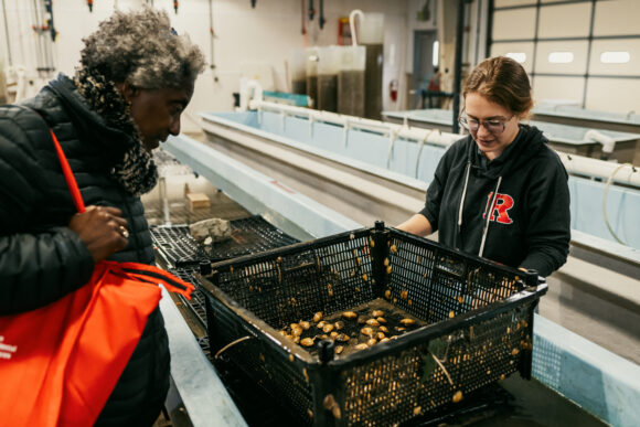 Two women standing on either side of a long open tank of water. Between them is a black crate of clams, seemingly fished out of the water. The woman on the left looks down at the crate as the woman on the right is mid-explanation.