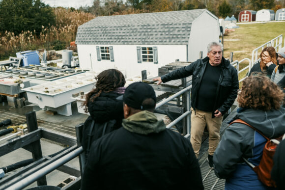 A group of people standing on a metal landing overlooking an open-air facility. One man is pointing down at the facility as he explains its purpose. 