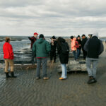 A group of people meandering around several rows of mesh bags elevated on rebar racks. While they mostly stand on sandy land, the waters creep in from the left.