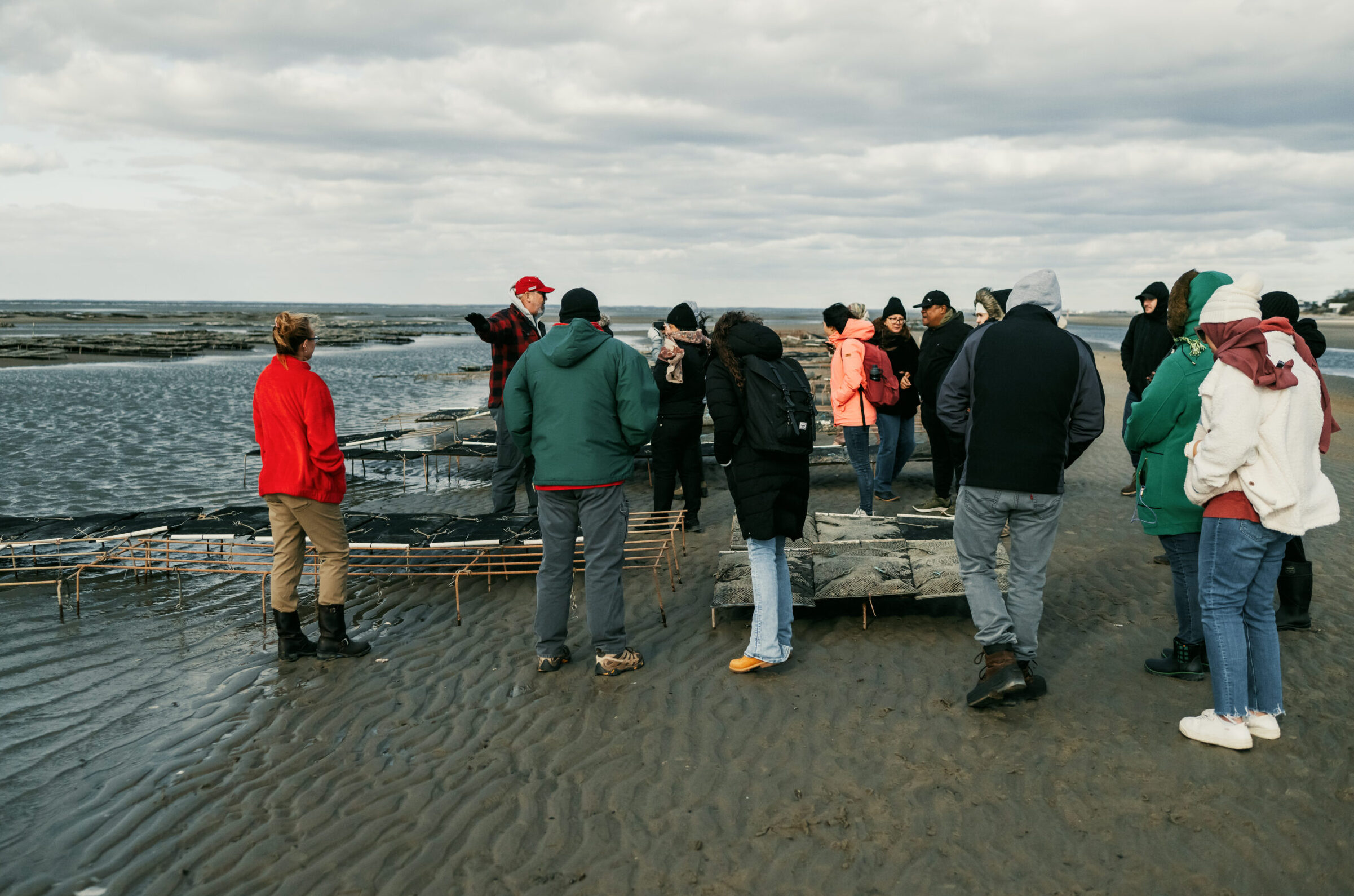 A group of people meandering around several rows of mesh bags elevated on rebar racks. While they mostly stand on sandy land, the waters creep in from the left.