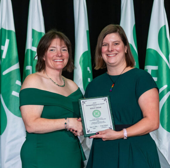 Two individuals on a stage at an awards ceremony standing in front of a backdrop of flags