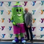 A mascot, representing a dill pickle, and a person stand in front of a banner