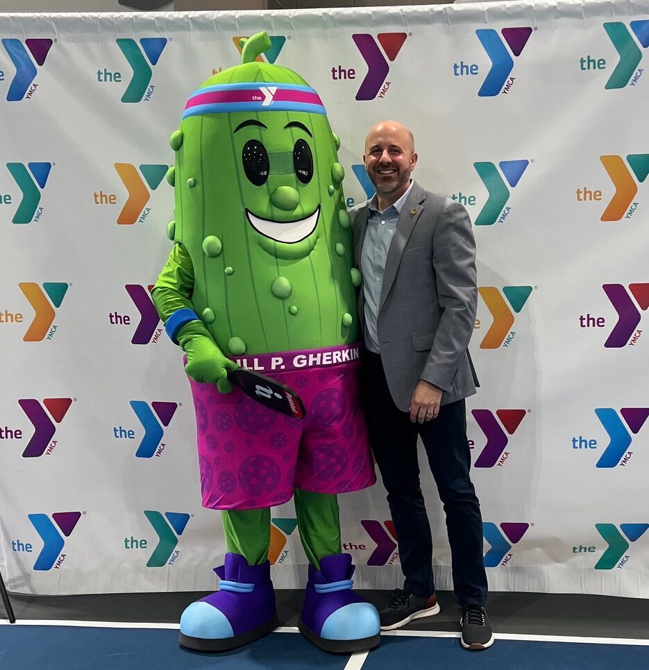A mascot, representing a dill pickle, and a person stand in front of a banner