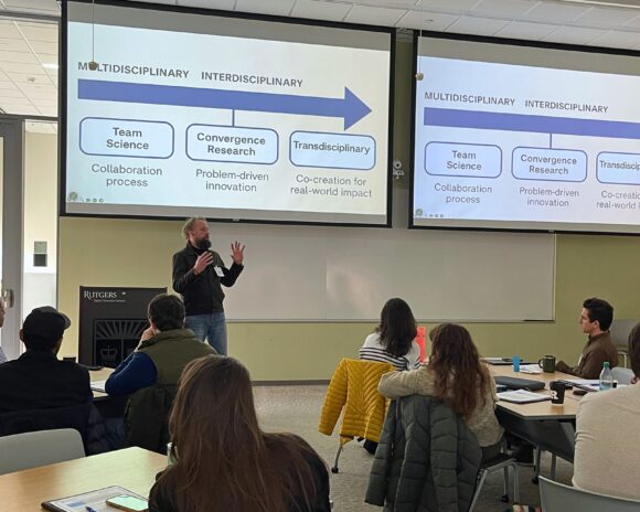 A man speaking to several attendees, all sat around different tables. Behind him, a slide depicting the relationship between multidisciplinary, interdisciplinary, transdisciplinary, team science, and convergence research is projected.
