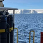 Sampling rosette with gray sampling bottles at left, the ship’s rail at lower right, and the face of the ice shelf in the background.