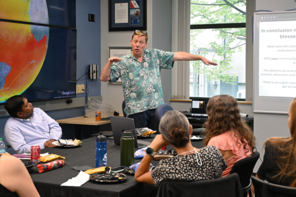 A person standing in front of a sat group mid-lunch. He is mid-explanation, Emphasizing a point by extending his left arm out to his side and his right arm in front of him.