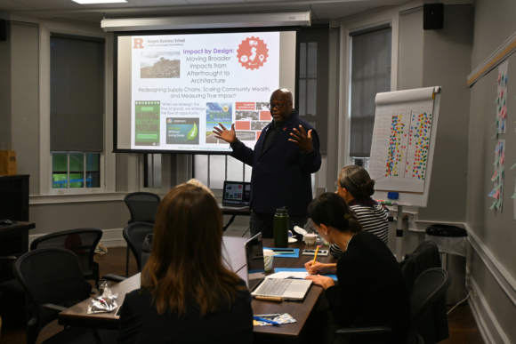 A person stands in front of a group taking notes at a table. He is mid-presenting, accompanied by a projected slide behind him titled “Impact by Design: Moving Broader Impacts from Afterthought to Architecture.”