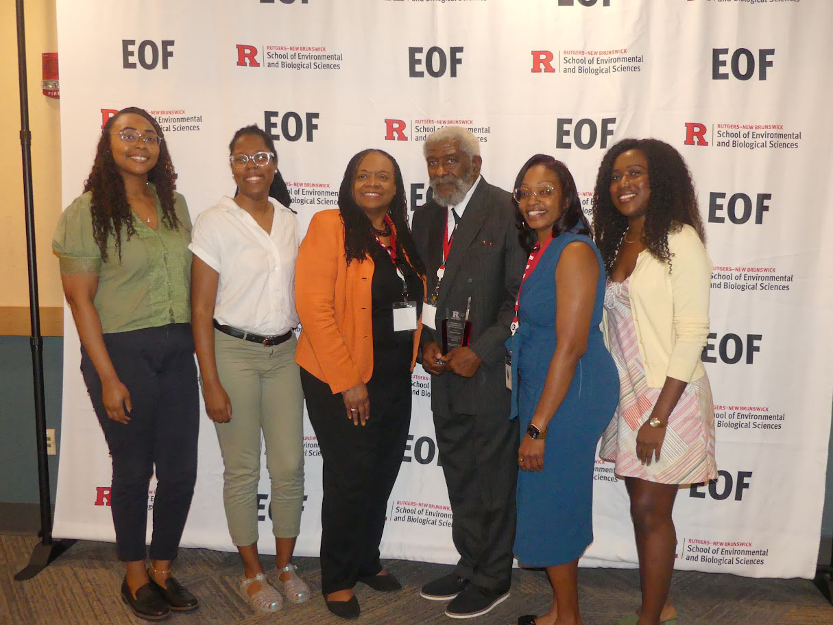 Six individuals representing alumni of a college readiness program are photographed standing in front of a banner with current and past leadership of the program.