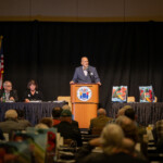 A person at a podium speaks to a crowd while a panel sits next to him
