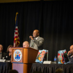A person at a podium speaks to a crowd while a panel sits next to him