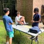 STEM Amb 2025 Newsroom Article - 30 A person talks to two others while sitting on a table